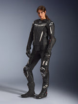 Woman wearing Alpinestars Stella GP-R7 2-Piece Leather Suit in Black, professional motorcycle racing gear featuring white logo details and accents, paired with matching black racing gloves and boots, studio full-body shot.