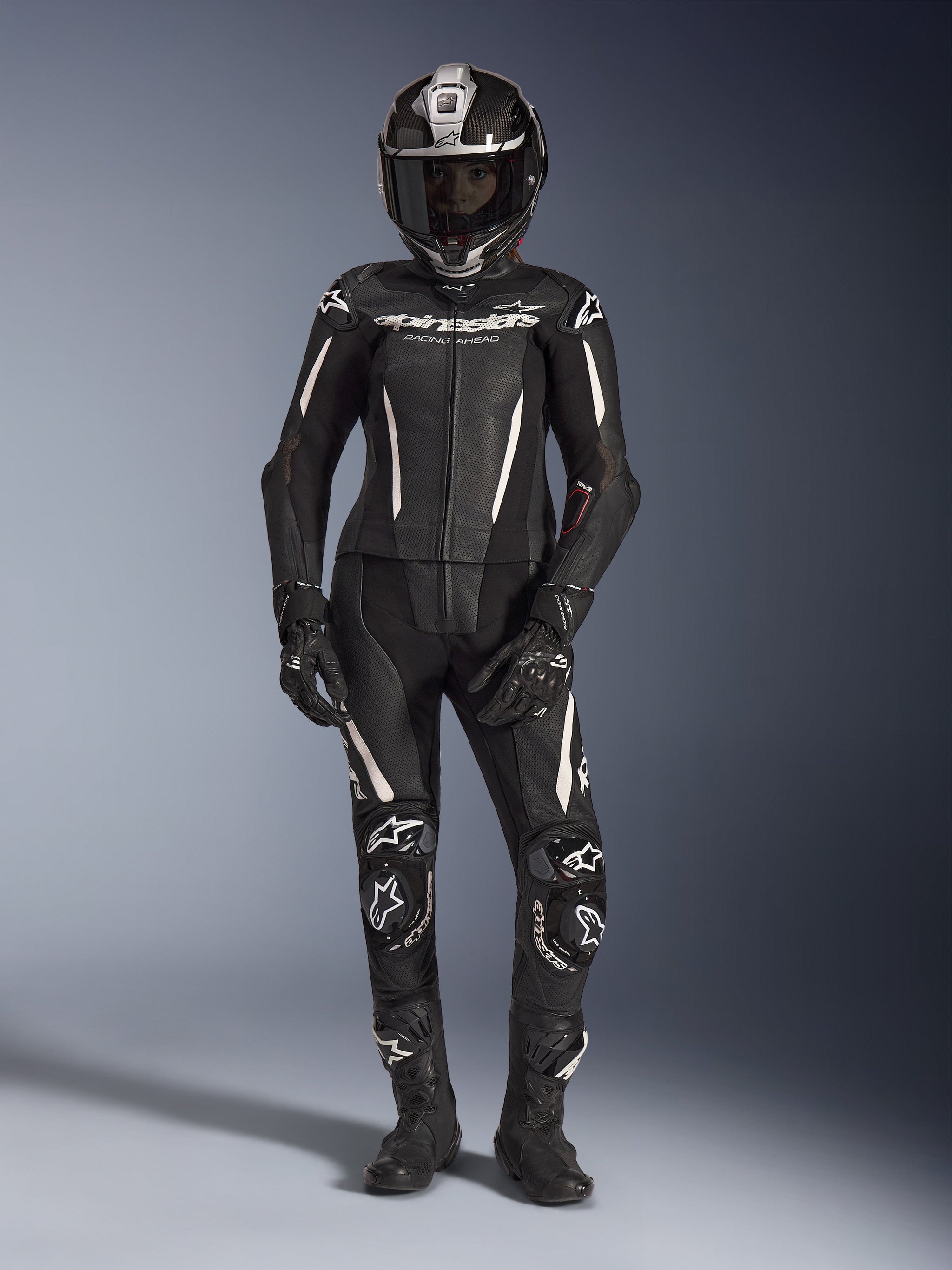 Woman rider wearing the Alpinestars Stella GP-R7 2-Piece Leather Suit in black with white accents, complete with a full-face carbon fiber helmet, black racing gloves, and boots, featuring perforated leather panels and integrated protective armor, professional motorcycle racing gear for track and street performance