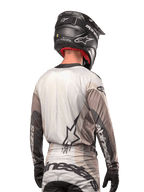 Motocross rider from behind wearing the Alpinestars 2024 Techstar Pneuma Jersey in Dark Sand Iron Dust Gray, paired with a black carbon-finish helmet, featuring a large star logo on the back and Alpinestars branding on the sleeves.